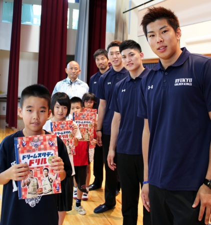 ▲沖縄市立室川小では、選手から子どもたちに直接贈呈された（写真右手前から、渡辺竜之佑選手、津山尚大選手、田代直希選手、金城茂之選手、安永淳一取締役と児童代表）