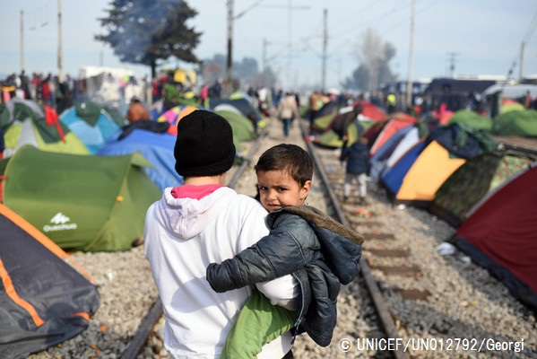 線路の周りに立てられたテントの間を、弟を抱いて歩く難民の少年。（ギリシャ）© UNICEF_UN012792_Georgiev