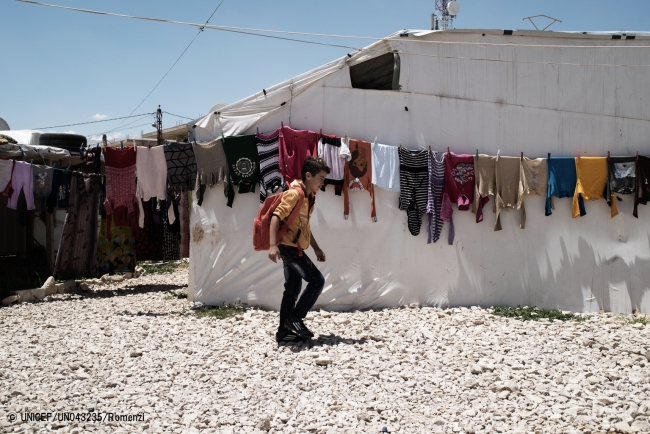 非公式居住地に干された洗濯物の列を通り過ぎながら、学校に通うシリア難民の男の子（レバノン、ベッカー高原）© UNICEF_UN043235_Romenzi